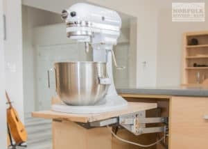 a natural-finish kitchen cabinet with a mechanical mixer lift holding a white kitchen aid mixer