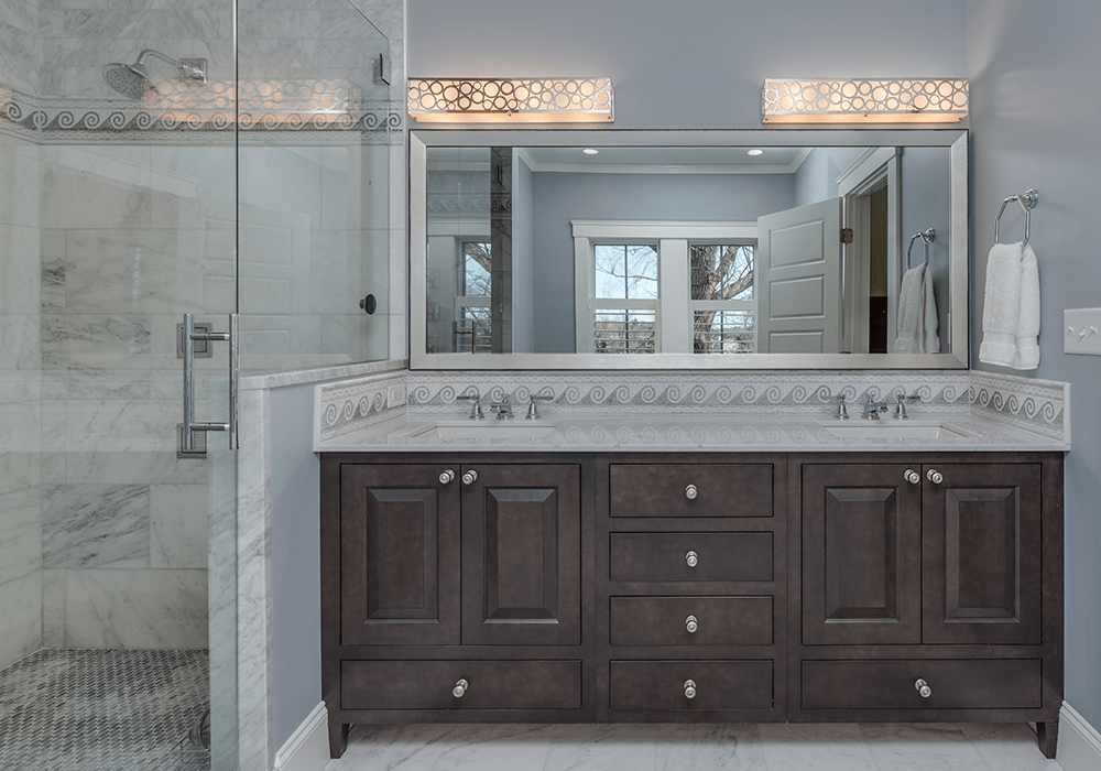 a custom Showplace Cabinetry bathroom vanity in a dark-stain finish with double sinks and a nautical backsplash next to a walk-in glass shower