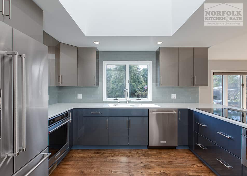 a modern, high-gloss kitchen with gray wall cabinets, blue base cabinets and stainless steel appliances with a kitchen window leading to the backyard