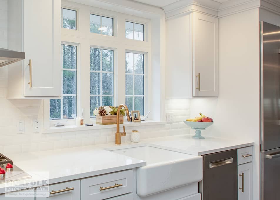 close up of a white apron sink and gold kitchen faucet in a white kitchen facing a large window