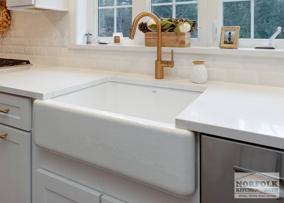 close up of a white apron sink in front of a large kitchen window with a gold kitchen faucet