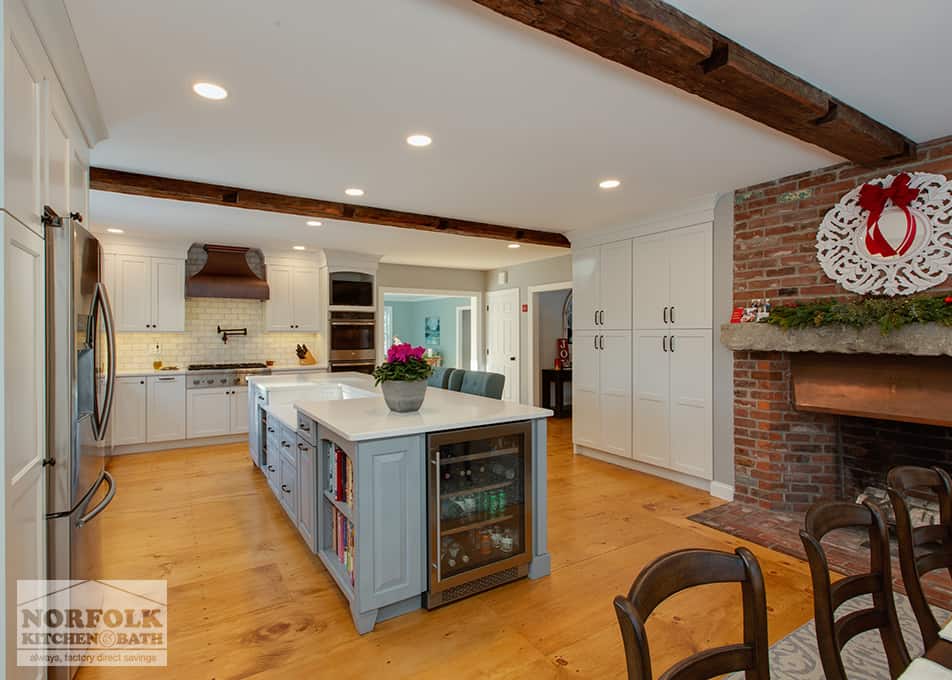 a large grey and white kitchen with a large furniture-style island, a wine fridge on the end of the island, pantry cabinets to the right with a brick fireplace next to them