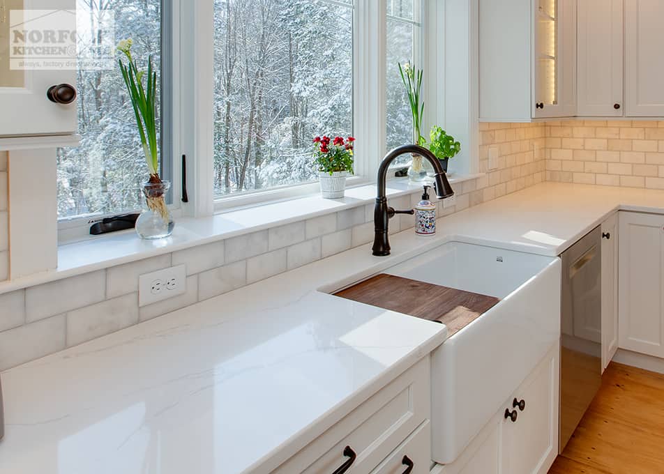 angled view of white stone countertops on white kitchen cabinets ending at a white apron sink with a pull down metal faucet in front of a large picture window